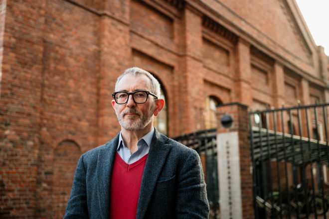 A man with grey hair and glasses, wearing a red sweater and dark blazer, stands outside in front of a large brick building