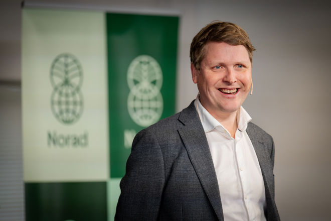 En mann i dress og hvit skjorte står foran en Norad-plakat innendørs.