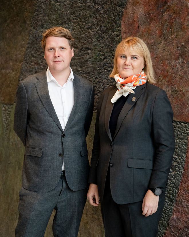 En mann og en kvinne står side om side, begge kledd i mørke dresser foran en steinvegg.