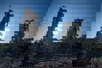 The photo shows two destroyed high-rise buildings standing among large amounts of rubble and debris. The sun shines low between the damaged structures, and the sky is clear and blue. The atmosphere is marked by silence and devastation, suggesting the area has suffered extensive damage.