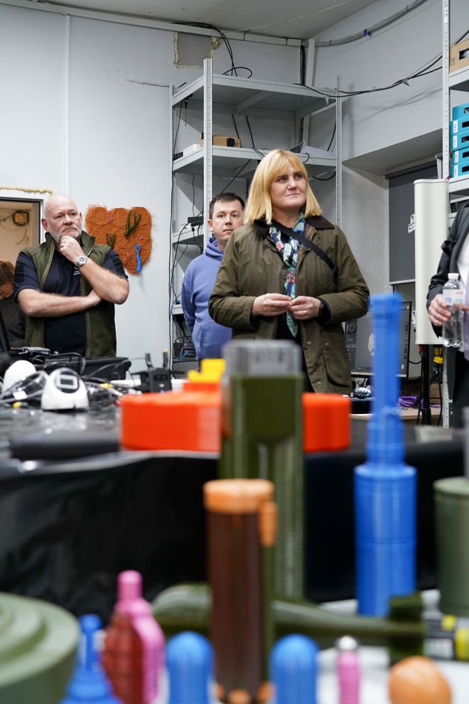 Several adults stand in a room with technical equipment and colorful models of explosives or ammunition displayed on a table in the foreground.