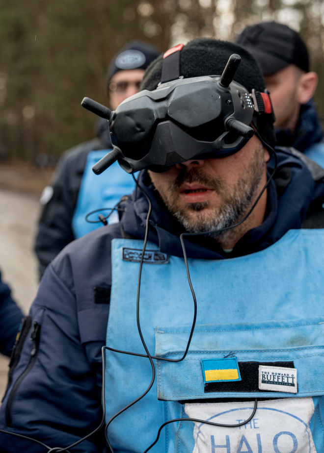 A person wearing protective gear and a blue vest with a Ukrainian flag and "The HALO Trust" logo stands outdoors, with another person in similar clothing in the background.