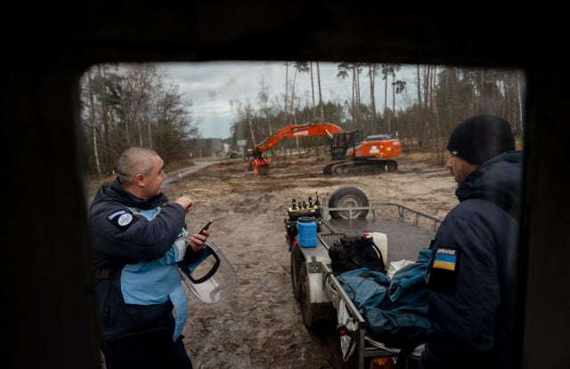 To menn i arbeidsklær står ved en tilhenger med utstyr på en gjørmete byggeplass, med en gravemaskin i bakgrunnen.