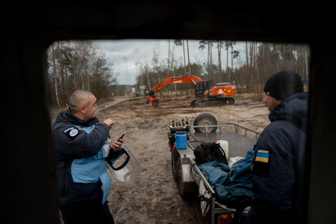Two men in uniforms stand next to a trailer with equipment on a muddy construction site, while an orange excavator operates in the background.