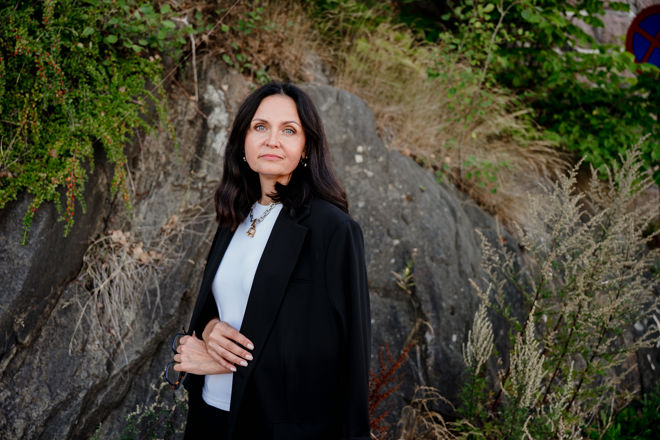 A woman with shoulder-length dark hair is standing outdoors in front of a rocky wall surrounded by greenery and wild plants. She is wearing a white top, a black blazer, and a necklace. The setting feels natural and calm, with plants and rocks forming the background.
