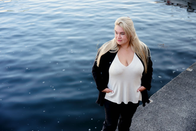 The image shows a woman with long blonde hair standing on the edge of a dock or waterfront. 