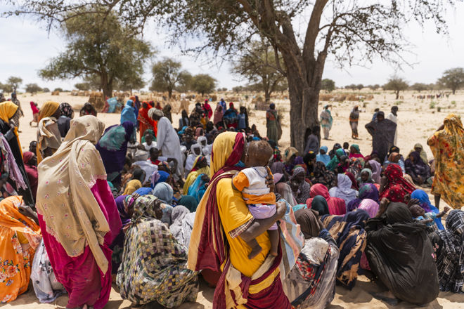A large group of women and children, many wearing colorful clothing, are gathered outdoors under the shade of trees in a dry, sandy area.