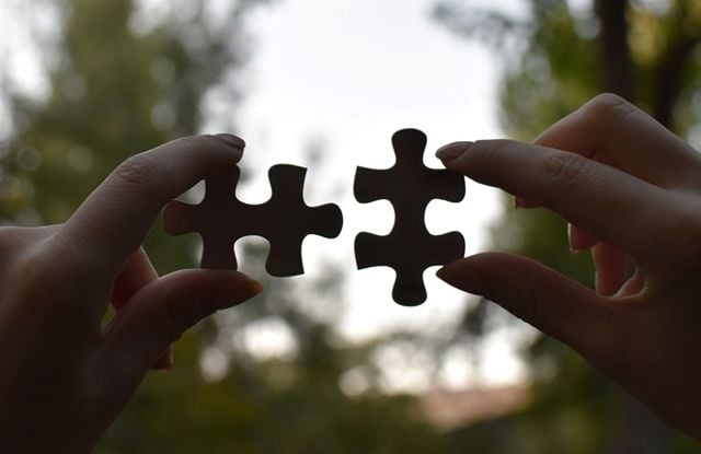 The image shows two hands, each holding a puzzle piece. The hands are facing each other, and the puzzle pieces are positioned so that they almost fit together, but are still slightly apart. 