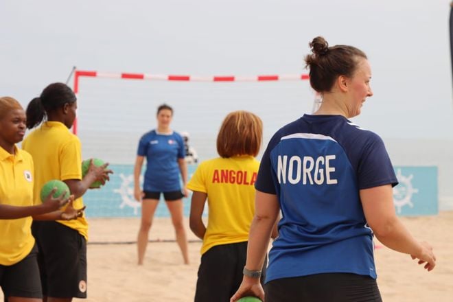 Flere personer deltar på en aktivitet eller trening på en sandbane ved sjøen. Noen har på seg gule t-skjorter med teksten "ANGOLA" på ryggen, mens én person har på seg en blå t-skjorte med teksten "NORGE" på ryggen. En håndball-lignende ball holdes av flere av deltakerne.