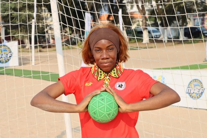 En kvinne står foran et mål på en sandvolleyballbane eller lignende uteområde, med en grønn ball holdt med begge hender foran brystet. Vedkommende har på seg en rød trøye med et emblem og mønstret krage, som gir inntrykk av å være en del av et lag eller organisasjon. Bakgrunnen viser nett, sand og noen reklameskilt, samt trær og biler i det fjerne. 