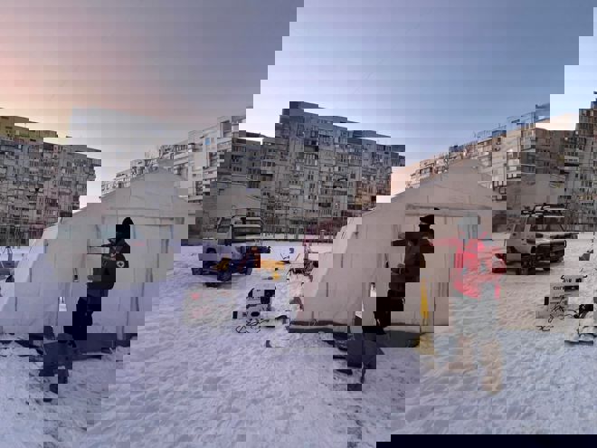 En ung mann i Røde Kors-uniform står utenfor et stort, lyst telt på en snødekt plass, med en firehjulstrekker og boligblokker i bakgrunnen