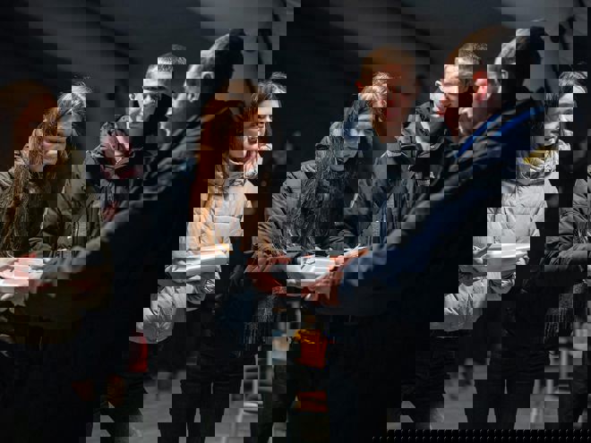 Several young people in jackets receive food containers from a person in uniform inside a tent.    Copy response