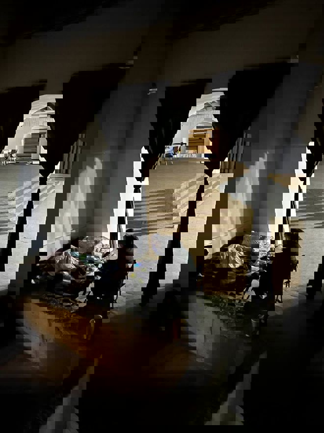 En person sitter ved siden av en feltseng med en sovende under tepper i et mørkt rom, mens et opplyst rom med stoler og hyller vises i bakgrunnen.