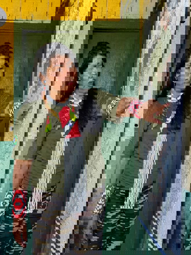 En kvinne står foran en vegg og peker på et stort kart som henger opp. Hun har langt, mørkt hår og bærer en grønn T-skjorte med et brasiliansk flagg på ermet, kamuflasjemønstrede bukser, og fargerike armbånd i rødt og hvitt. 