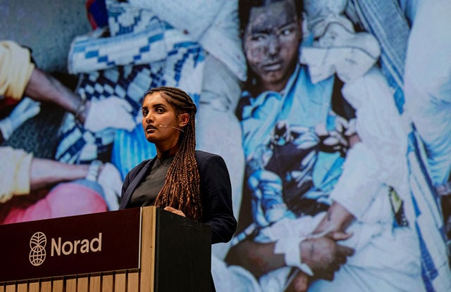 A young woman stands behind a podium with the Norad logo, in front of a large screen displaying an image of people in the background.