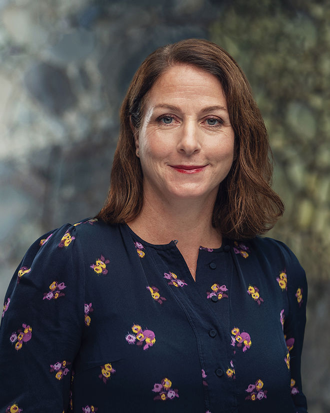 En kvinne med brunt, skulderlangt hår har på seg en mørk bluse med blomster, og står foran en uskarp bakgrunn.